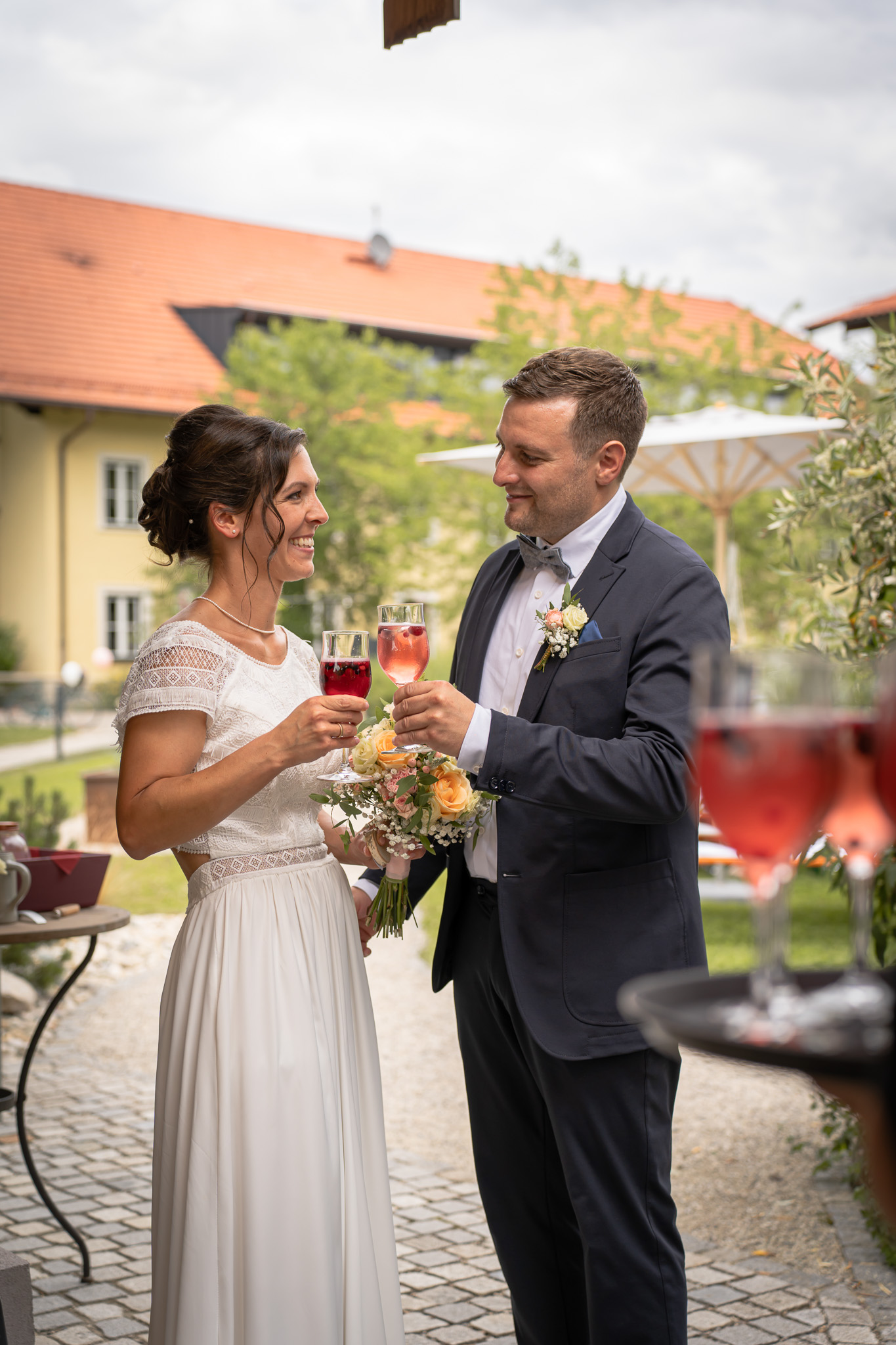 Authentischer Sektempfang mit Brautpaar – Hochzeitsfotografie | Lindl Photography Brautpaar stößt beim Sektempfang miteinander an und lacht.