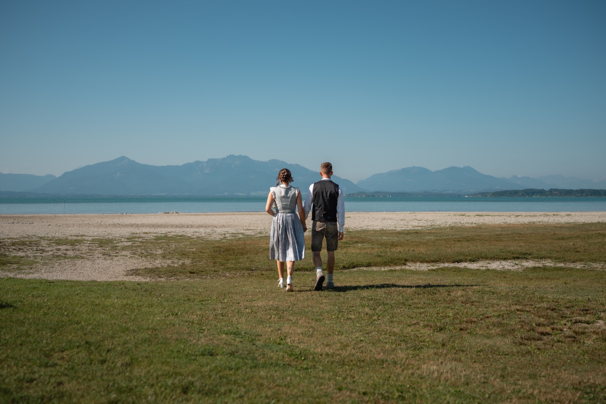 Brautpaar in Tracht spaziert am Chiemsee entlang, die Berge im Hintergrund.