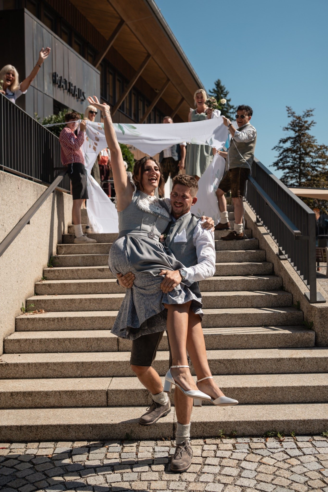 Standesamtliche Hochzeit: Bräutigam trägt seine Braut auf einer Treppe, während Gäste mitfeiern.